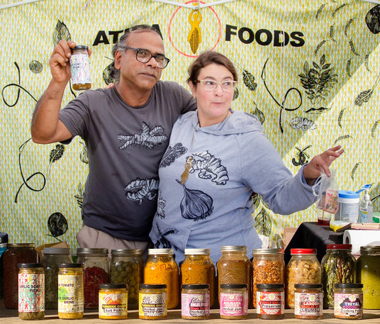 Two people standing behind a display of jars with 'Atlin Foods' branding on a decorative wall.