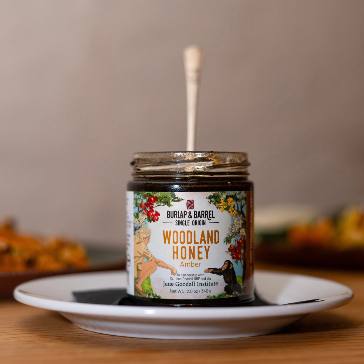 A jar of Burlap & Barrel Single Origin Woodland Honey Amber with a wooden honey dipper resting inside, displayed on a white ceramic plate on a wooden table with a blurred background.