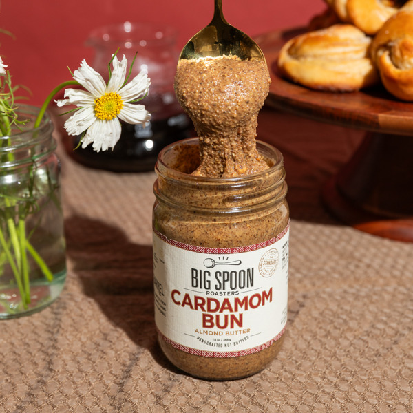 Jar of 'Big Spoon Roasters Cardamom Bun Almond Butter' with a spoonful on top, set against a rustic background.