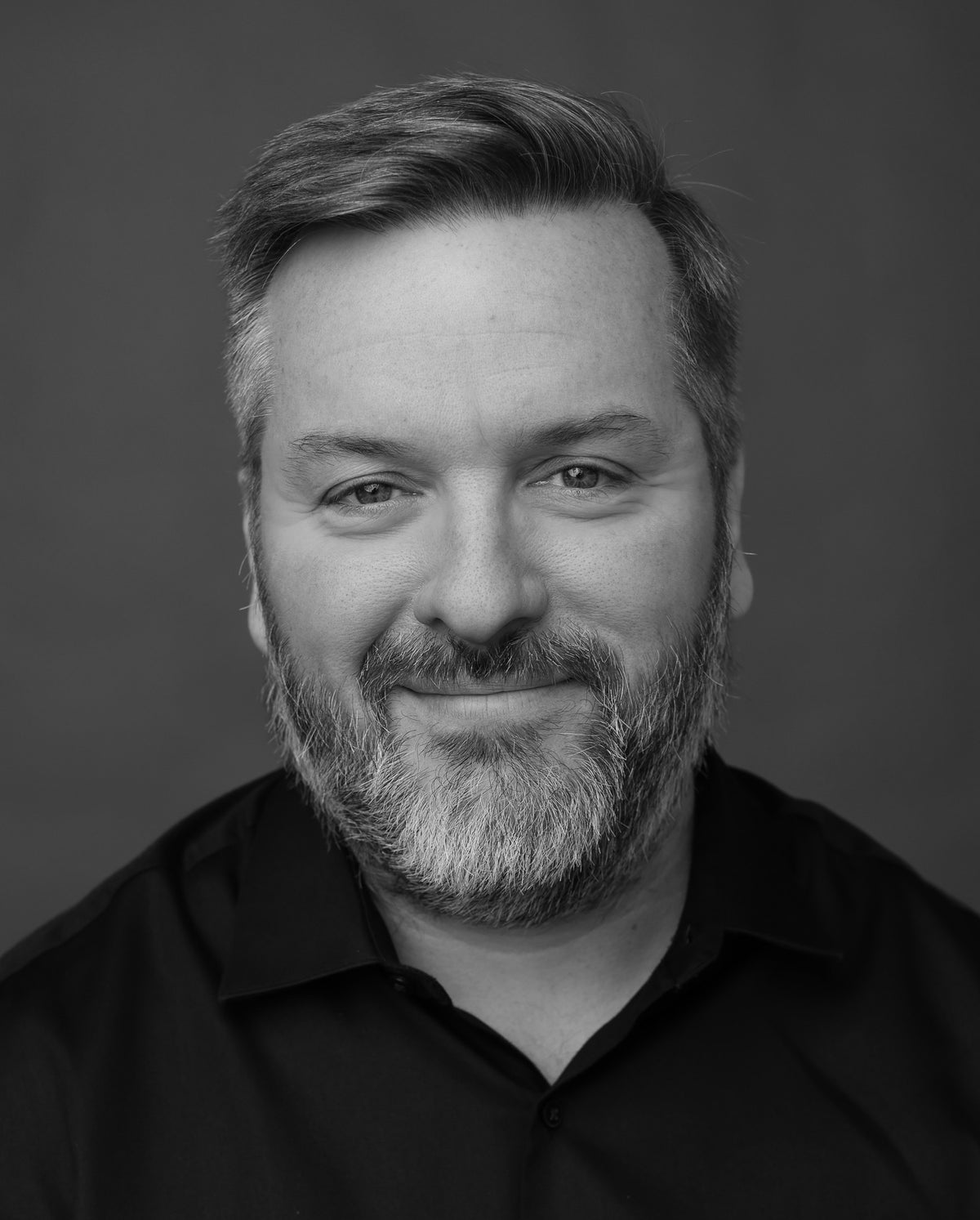A photo of a man with a beard smiling in front of a grey background.