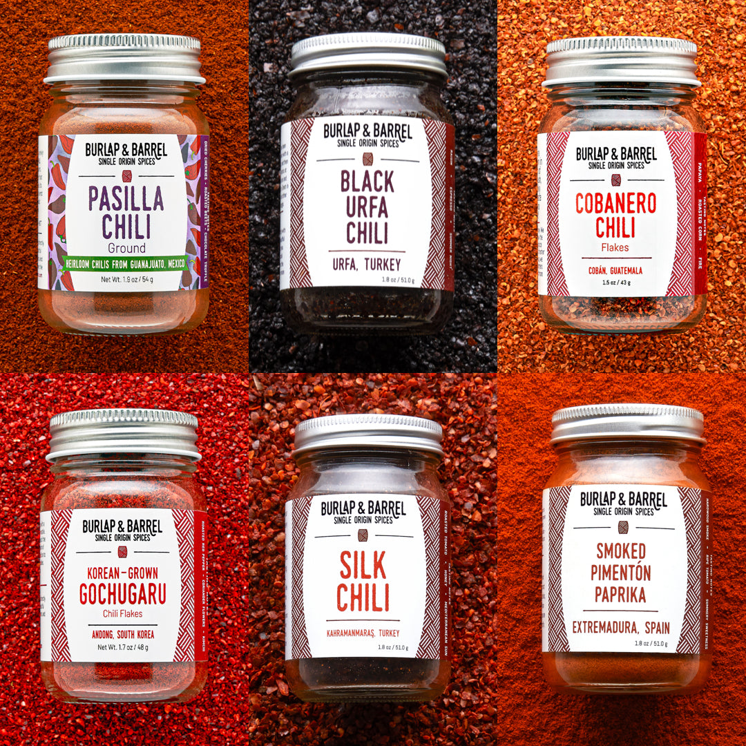 Collection of Burlap & Barrel chili and spice jars on a textured red and brown background.