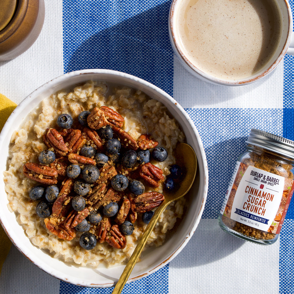 Bowl of oatmeal with blueberries and pecans, accompanied by a jar of cinnamon sugar crunch by Burlap & Barrel on a checkered tablecloth.