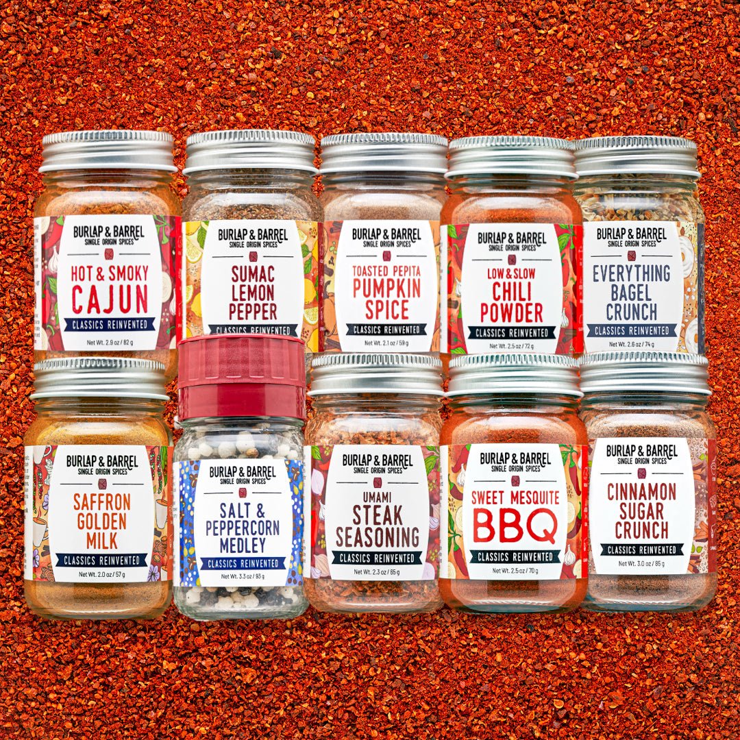 Eight glass spice jars from Burlap & Barrel arranged in two rows on a textured orange-red spice background. The labels show different spice flavors including Cajun, Lemon Pepper, Pumpkin Spice, Chili Powder, Everything Bagel Crunch, Saffron Golden Milk, Salt & Peppercorn Medley, Umami Steak Seasoning, Sweet Mesquite BBQ, and Cinnamon Sugar Crunch.