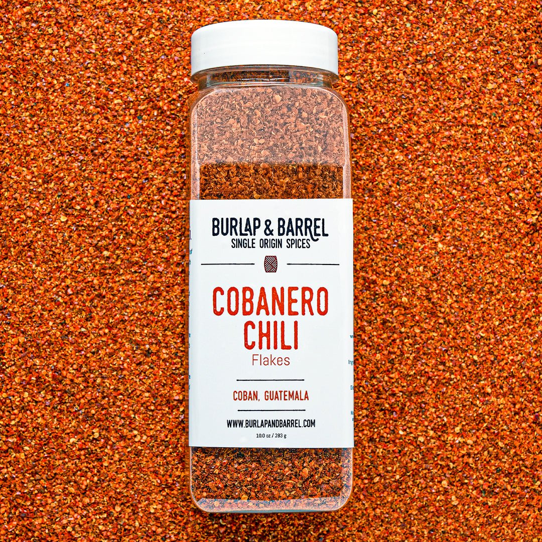 Container of cobanero chili flakes from burlap & barrel on a background of loose chili flakes.