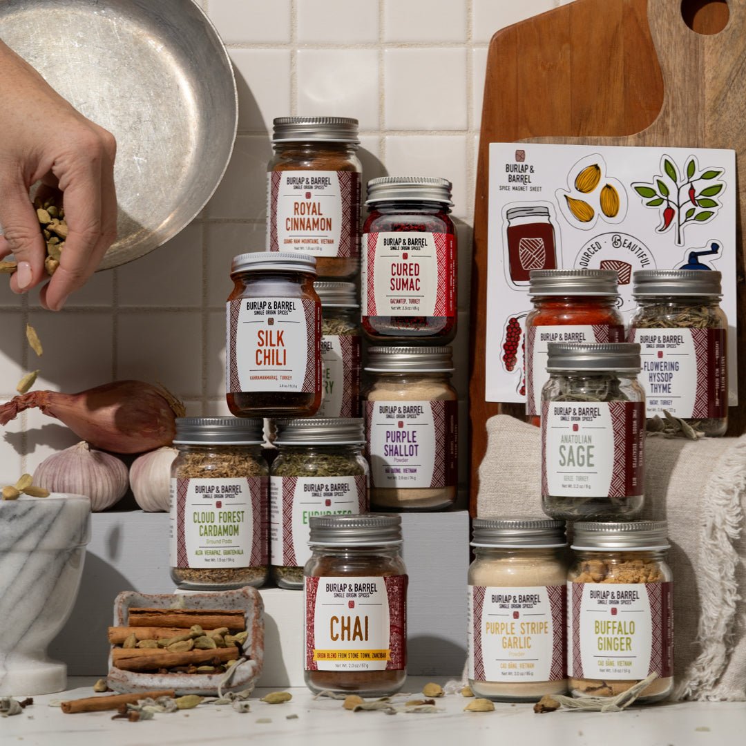 Jars of Burlap & Barrel spices and seasonings on a kitchen counter with a person preparing food.