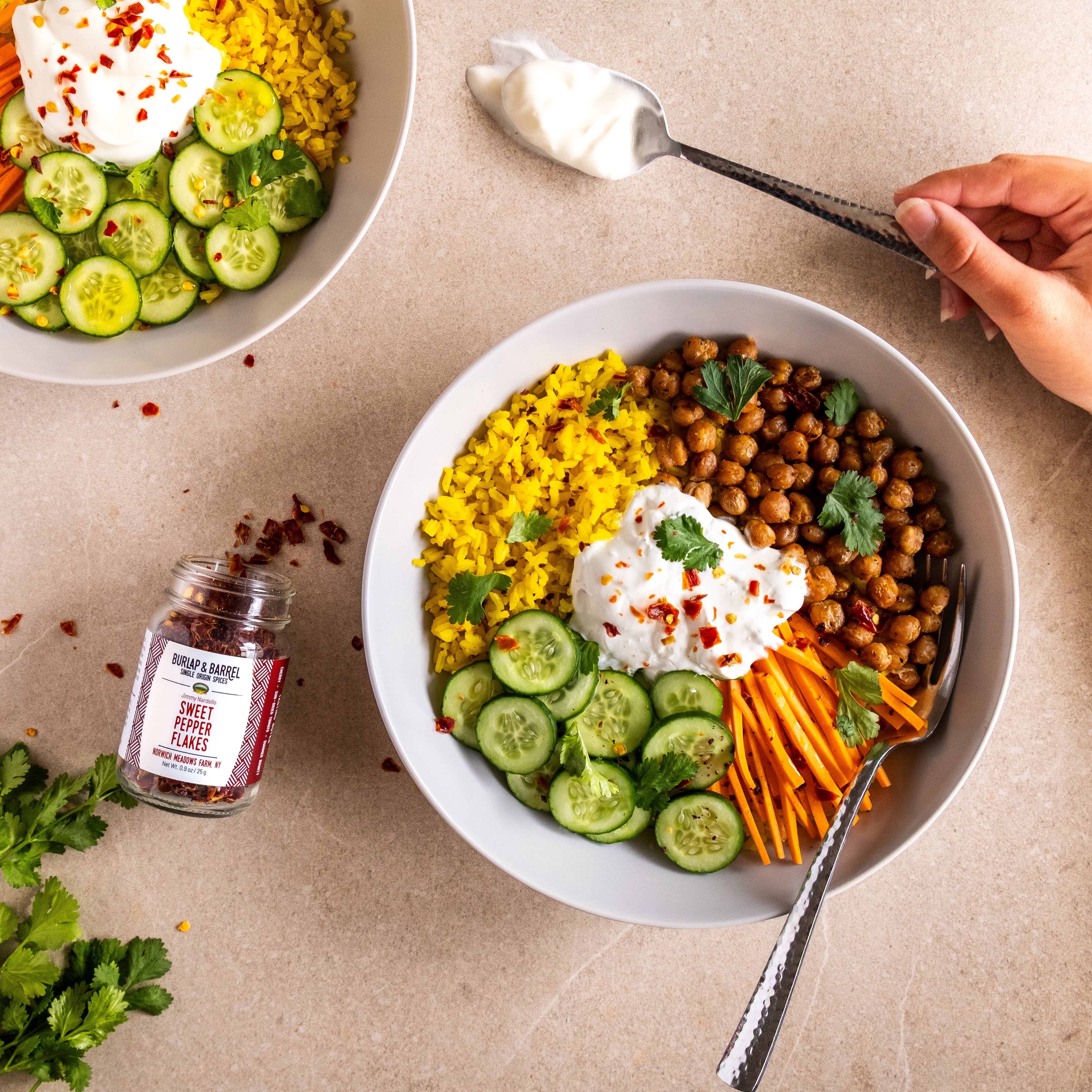Bowl of salad with chickpeas, carrots, and cucumbers on a beige surface with a jar Jimmy Nardello Sweet Pepper Flakes from Burlap & Barrel