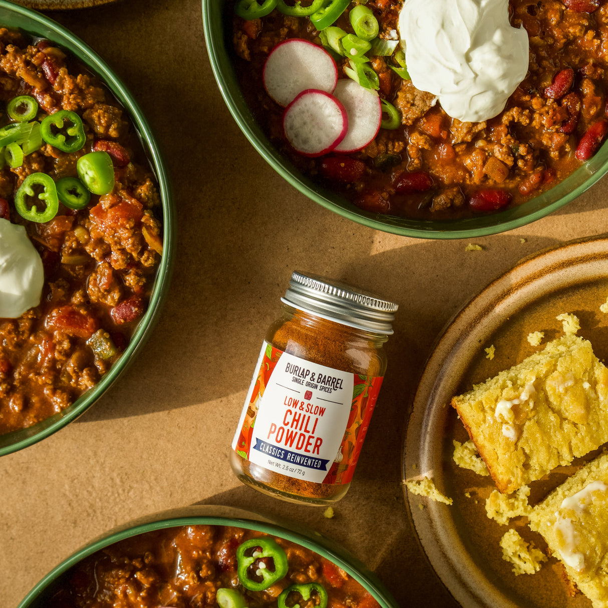 Bowls of chili with garnishes and a jar of Low & Slow Chili Powder from Burlap & Barrel
