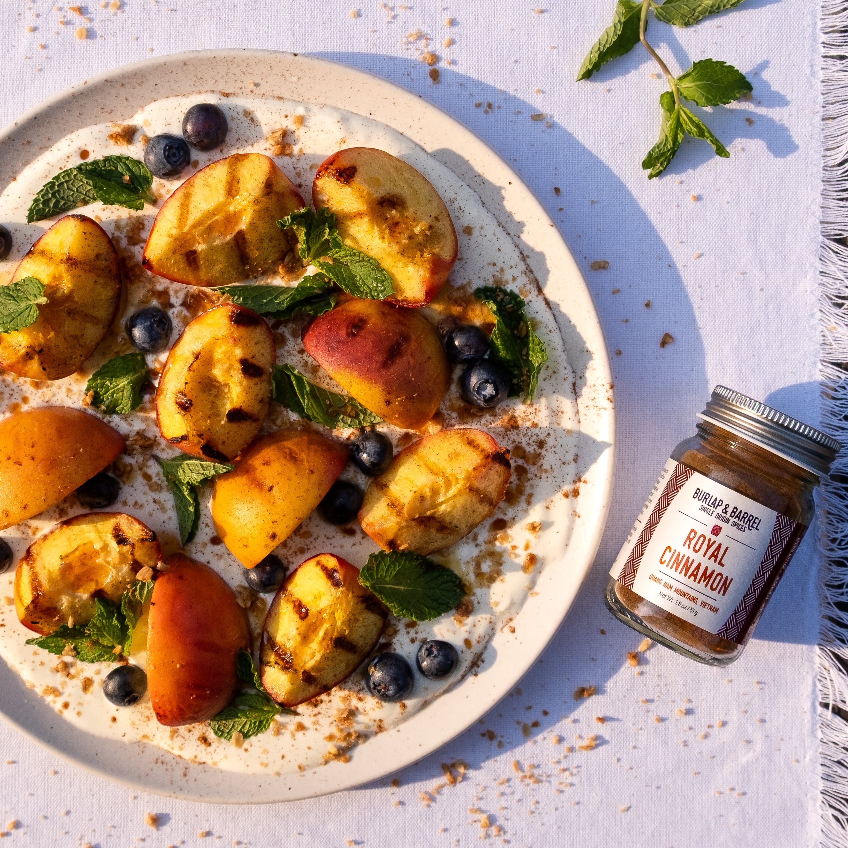 Plate of grilled peaches, blueberries, and mint leaves, accompanied by a jar of Royal Cinnamon from Burlap & Barrel