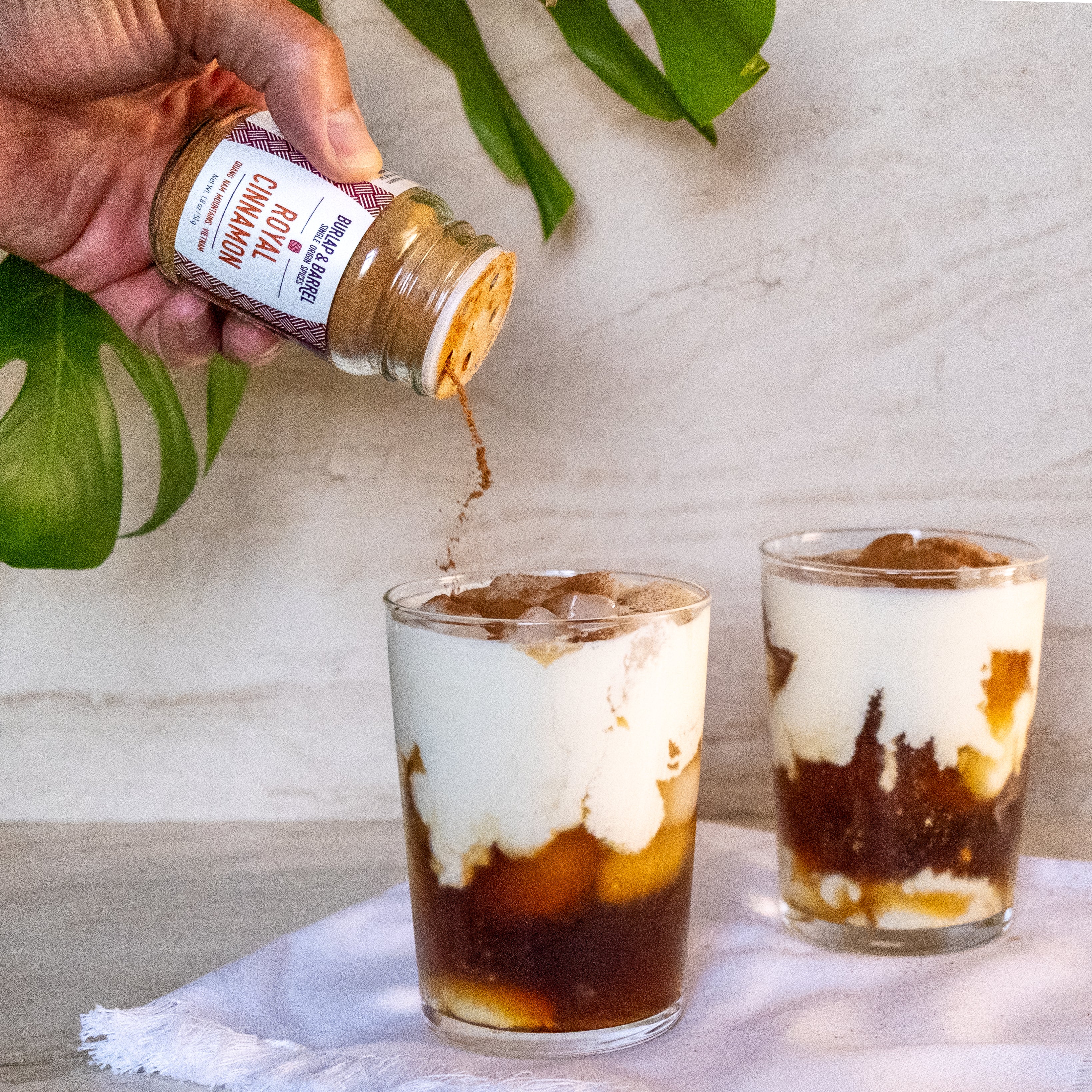 Hand pouring a jar of Royal Cinnamon into two glasses of iced coffee from Burlap & Barrel