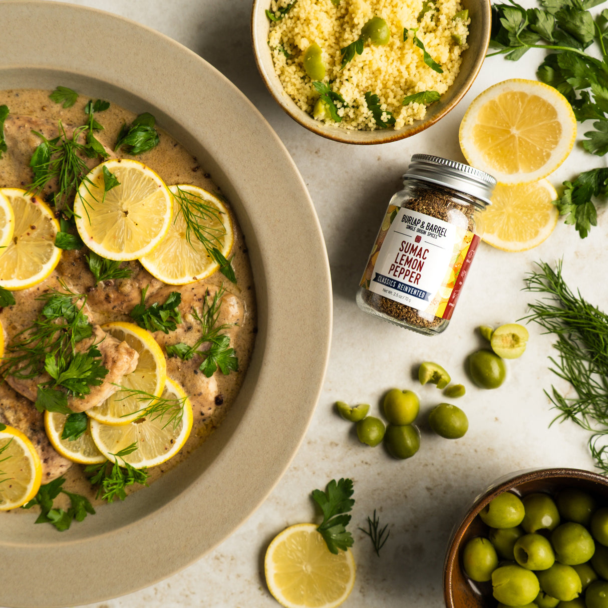 A jar of Sumac Lemon Pepper from Burlap & Barrel arranged next to a place of creamy chicken and lemon slices and olives.