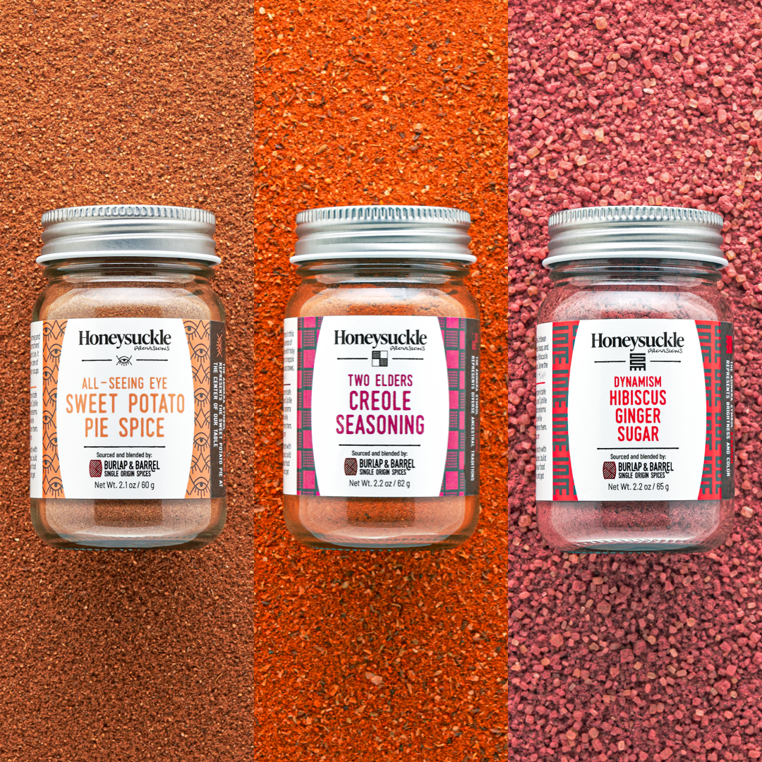 Three glass jars of Honeysuckle brand spice blends, arranged on a textured red background. The jars are labeled as 'SWEET POTATO PIE SPICE,' 'CREOLE SEASONING,' and 'HIBISCUS GINGER SUGAR.