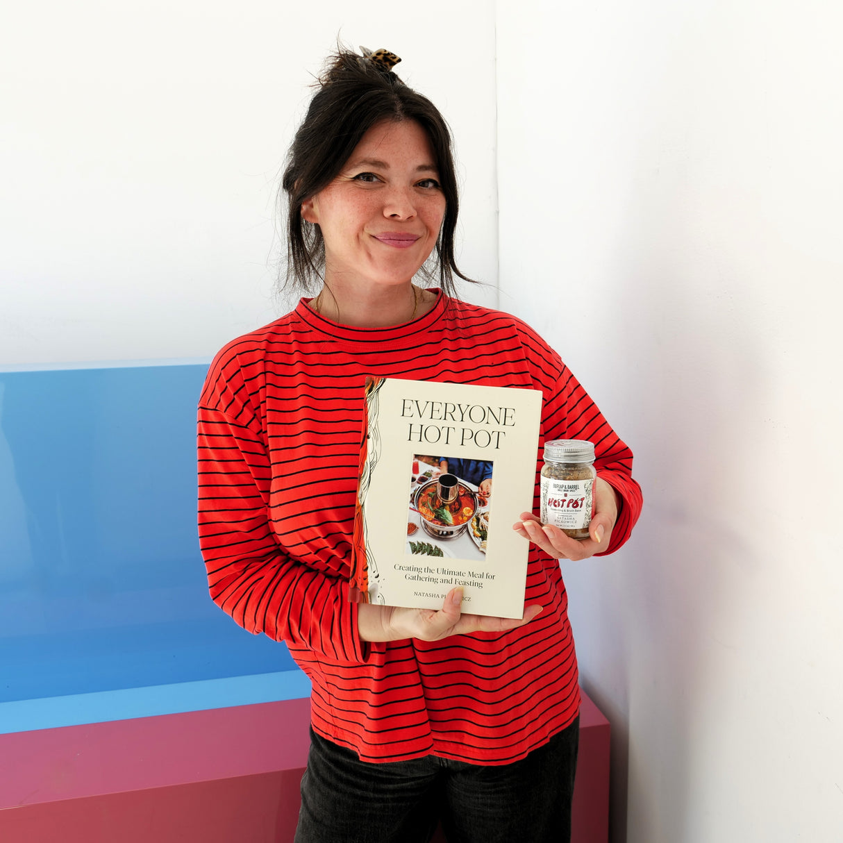 Photo of Natasha Pickowicz holding her cookbook and a jar of Hot Pot spice blend, by Burlap & Barrel