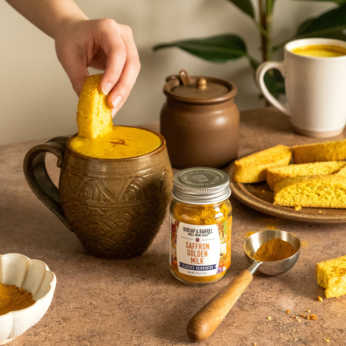 Person dipping a piece of biscotti into a mug with a latte made with Burlap & Barrel's Saffron Golden Milk and biscotti pieces on a table.