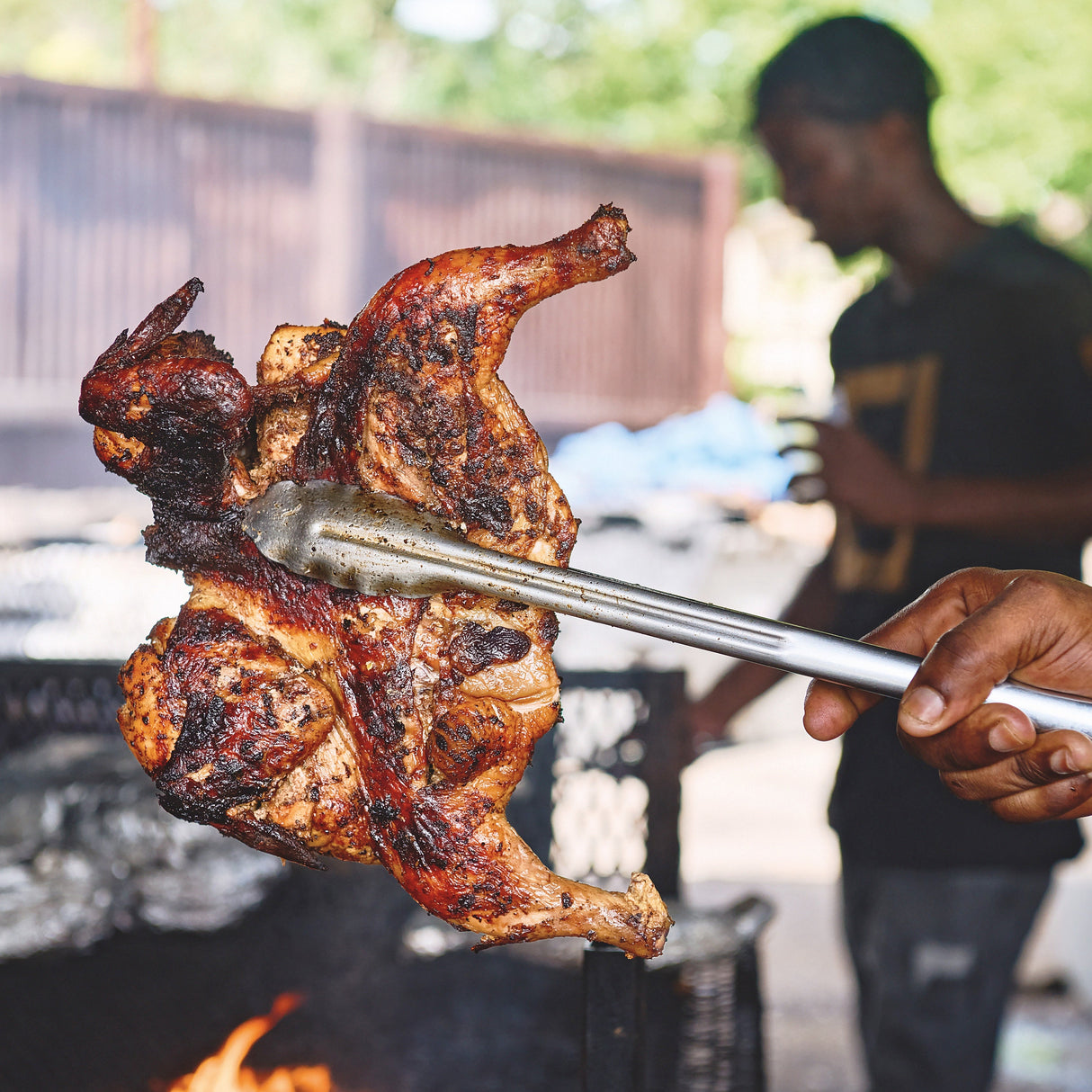 Person grilling a roasted chicken on an outdoor grill with trees in the background