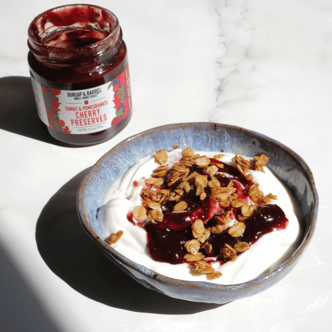 Bowl of yogurt with granola and cherry preserves next to a jar of cherry preserves on a marble surface.