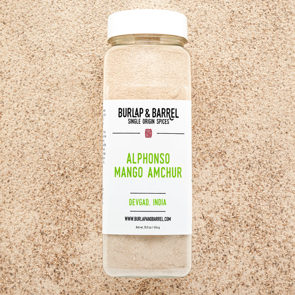 A jar of Alphonso Mango Amchur spice on a background of loose amchur with the label 'BURLAP & BARREL' and 'Alphonso Mango Amchur