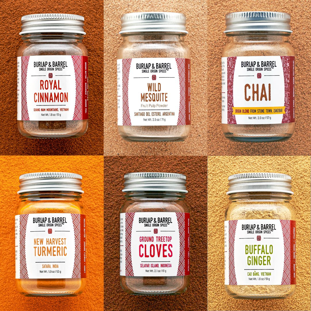Six spice jars arranged in a 2x3 grid on a textured warm background, each with a silver metal lid and a red-and-white label. The jars are labeled Royal Cinnamon, Wild Mesquite, Chai, New Harvest Turmeric, Ground Treetop Cloves, and Buffalo Ginger.
