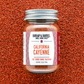 A glass jar of Burlap & Barrel California Cayenne spice blend, filled with orange-red seasoning and topped with a silver metal lid, set against a textured background of loose orange-red cayenne pepper.