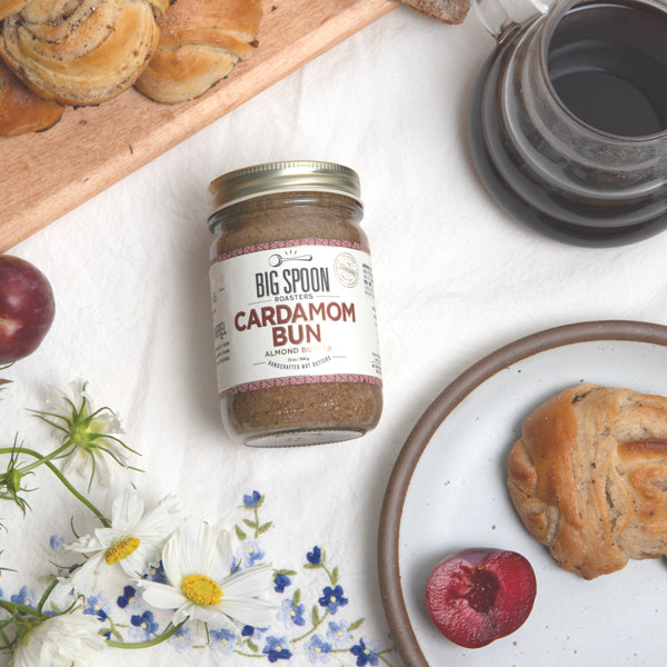 Jar of 'Big Spoon' Cardamom Bun spread on a table with pastries and flowers.