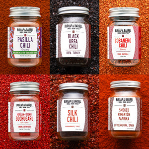Collection of Burlap & Barrel chili and spice jars on a textured red and brown background.