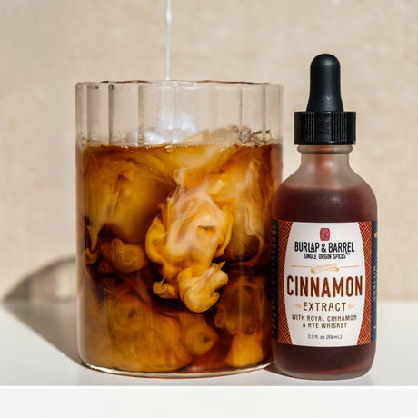 Glass cup of coffee with milk being poured into it next to a bottle with a dropper top of Burlap & Barrel Cinnamon extract on a tan background and white countertop.