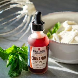 Dropper bottle of Burlap & Barrel cinnamon extract next to a spring of mint with a bowl of whipped cream and whisk in the background.  