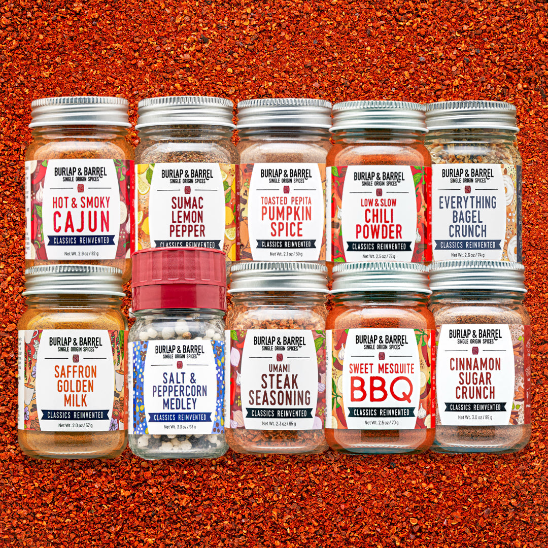 Eight glass spice jars from Burlap & Barrel arranged in two rows on a textured orange-red spice background. The labels show different spice flavors including Cajun, Lemon Pepper, Pumpkin Spice, Chili Powder, Everything Bagel Crunch, Saffron Golden Milk, Salt & Peppercorn Medley, Umami Steak Seasoning, Sweet Mesquite BBQ, and Cinnamon Sugar Crunch.