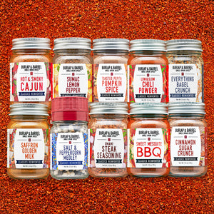 Eight glass spice jars from Burlap & Barrel arranged in two rows on a textured orange-red spice background. The labels show different spice flavors including Cajun, Lemon Pepper, Pumpkin Spice, Chili Powder, Everything Bagel Crunch, Saffron Golden Milk, Salt & Peppercorn Medley, Umami Steak Seasoning, Sweet Mesquite BBQ, and Cinnamon Sugar Crunch.