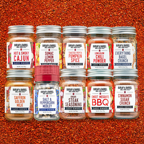 Eight glass spice jars from Burlap & Barrel arranged in two rows on a textured orange-red spice background. The labels show different spice flavors including Cajun, Lemon Pepper, Pumpkin Spice, Chili Powder, Everything Bagel Crunch, Saffron Golden Milk, Salt & Peppercorn Medley, Umami Steak Seasoning, Sweet Mesquite BBQ, and Cinnamon Sugar Crunch.