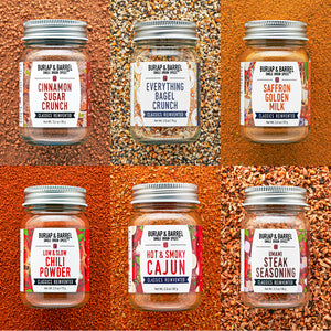 Six glass spice jars with silver lids arranged in a 2x3 grid, each set against a different spice background. The jars are labeled Burlap & Barrel, including Cinnamon Sugar Crunch, Everything Bagel Crunch, Saffron Golden Milk, Low & Slow Chili Powder, Hot & Smoky Cajun, and Umami Steak Seasoning.