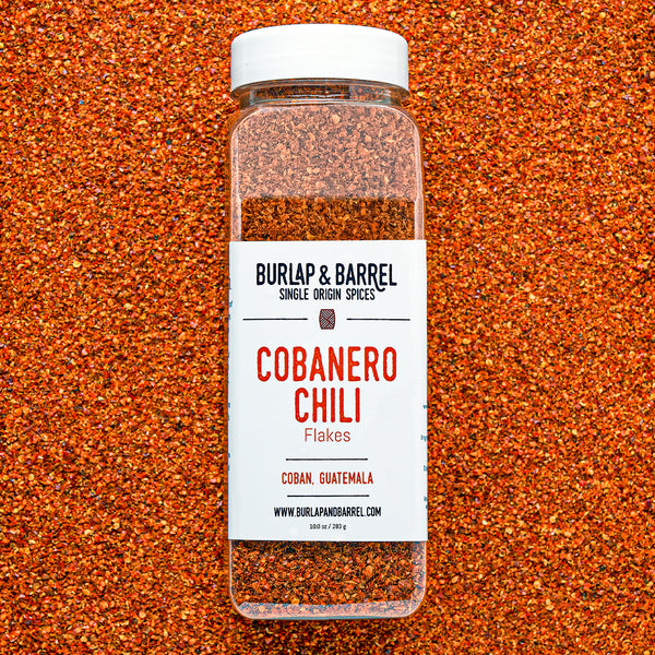Container of cobanero chili flakes from burlap & barrel on a background of loose chili flakes.
