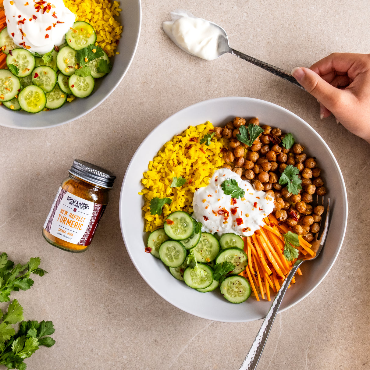 Bowl of salad with chickpeas, carrots, and cucumbers on a beige surface with a jar New Harvest Turmeric from Burlap & Barrel