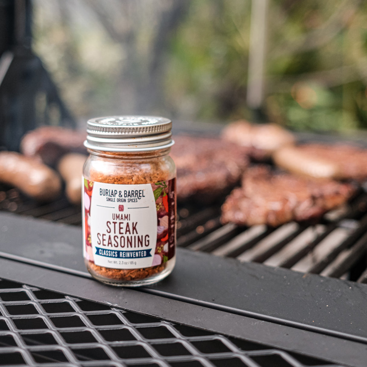 Jar of Burlap & Barrel Umami Steak Seasoning on a grill with cooked steaks in the background