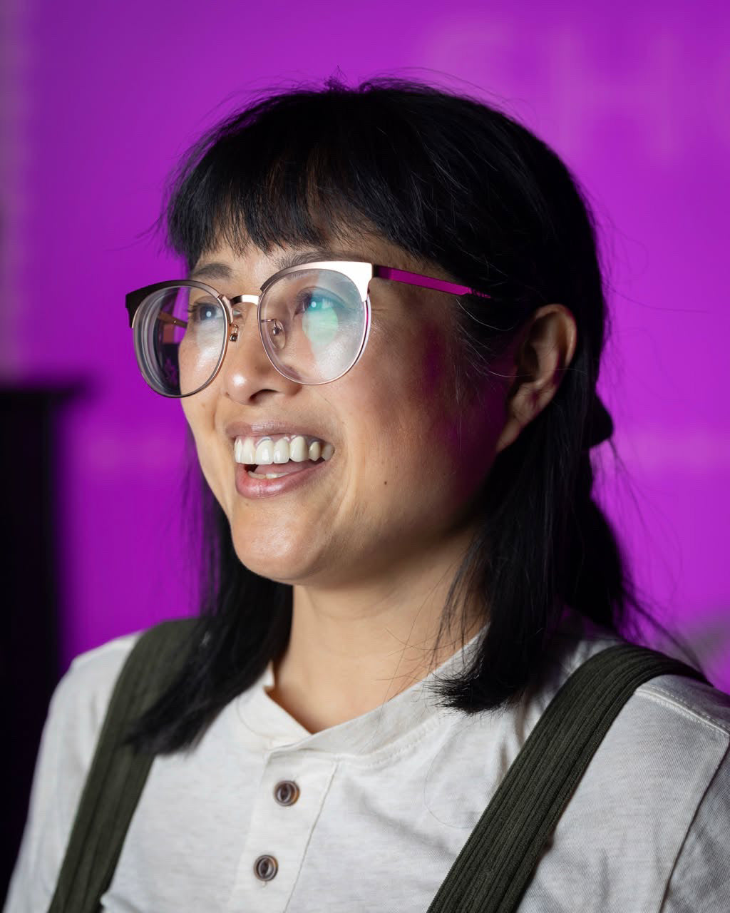 A profile of a woman with glasses smiling  in front of a bright magenta background.