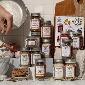 A styled kitchen scene showing multiple glass spice jars from Rupa & Barrel arranged on a countertop, with a hand pouring spices into a bowl at left and a wooden cutting board with spice illustrations at right.