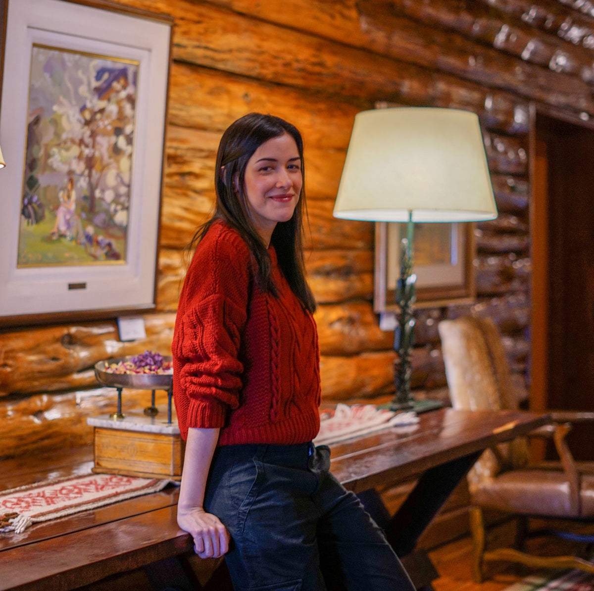 A photo of a woman wearing a reddish-orange sweater leaning on a desk with lamp in a log cabin-style room. 