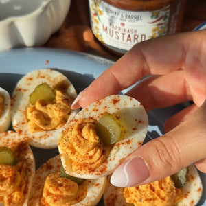 A photo of a hand holding a deviled egg topped with a pickle slice, with multiple deviled eggs and a Paprikash Mustard jar in the background. 