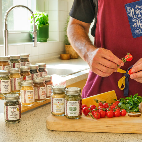 A person in a maroon apron stands at a kitchen counter preparing food while several jars of Burlap & Barrel spices are arranged on the counter, including garlic, rosemary, and peppercorn.