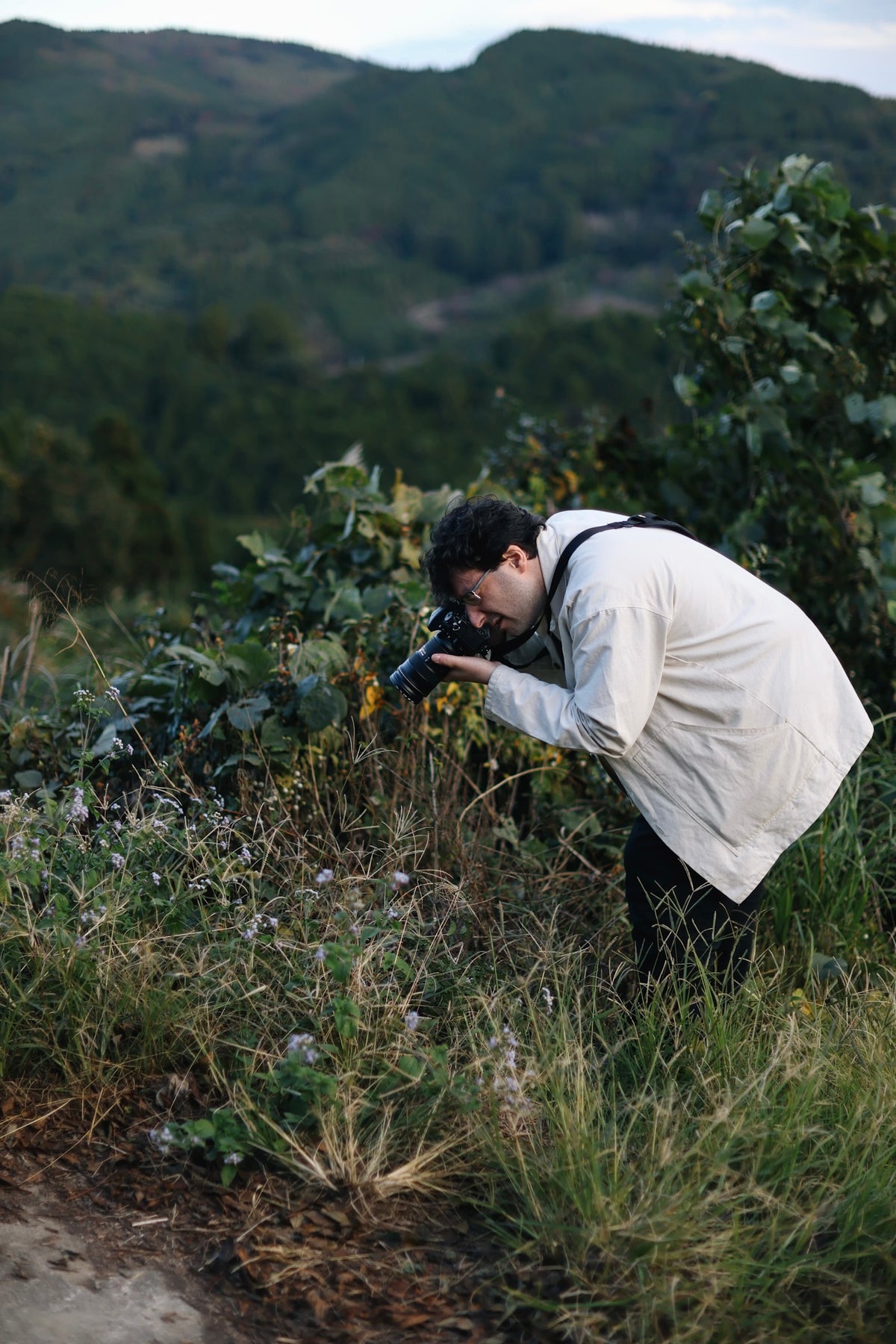 A man in a light-colored jacket and dark pants bends down in a grassy field, holding a camera to take a photograph of a lush green mountain landscape in the background.