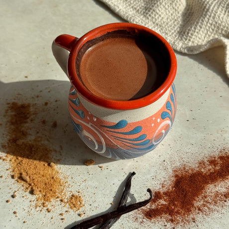 A hand-painted ceramic mug filled with a brown beverage, styled on a light surface with vanilla beans, cinnamon powder, and a textured cloth in the background.