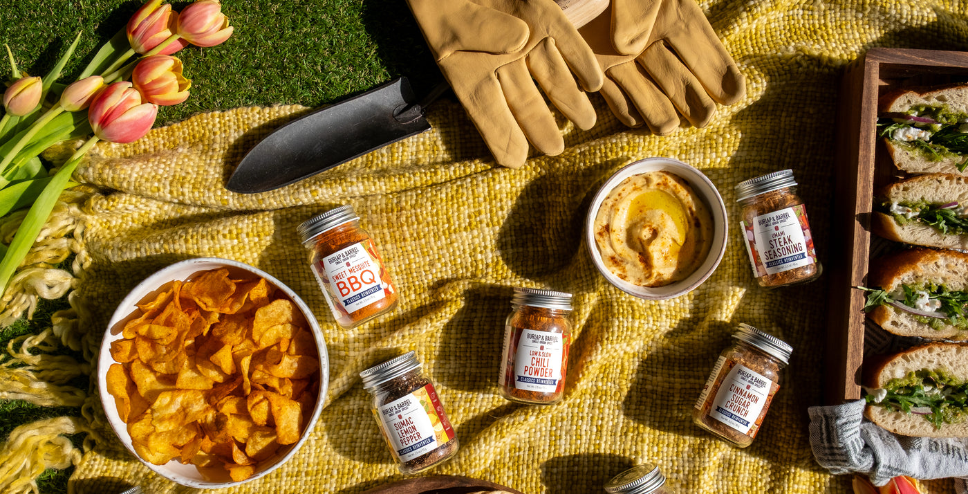 Outdoor picnic setup with chips, pickles, Burlap & Barrel spice jars and sandwiches on a yellow blanket.