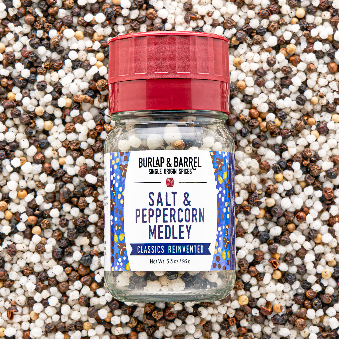 A bottle of Burlap & Barrel Salt & Peppercorn Medley spices, featuring a red cap and a white label with blue patterns, set against a background of mixed whole peppercorns.