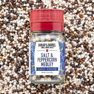 A bottle of Burlap & Barrel Salt & Peppercorn Medley spices, featuring a red cap and a white label with blue patterns, set against a background of mixed whole peppercorns.