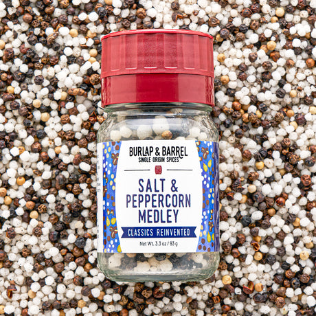 A bottle of Burlap & Barrel Salt & Peppercorn Medley spices, featuring a red cap and a white label with blue patterns, set against a background of mixed whole peppercorns.