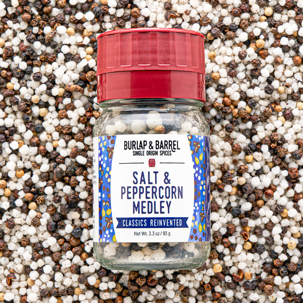 A bottle of Burlap & Barrel Salt & Peppercorn Medley spices, featuring a red cap and a white label with blue patterns, set against a background of mixed whole peppercorns.