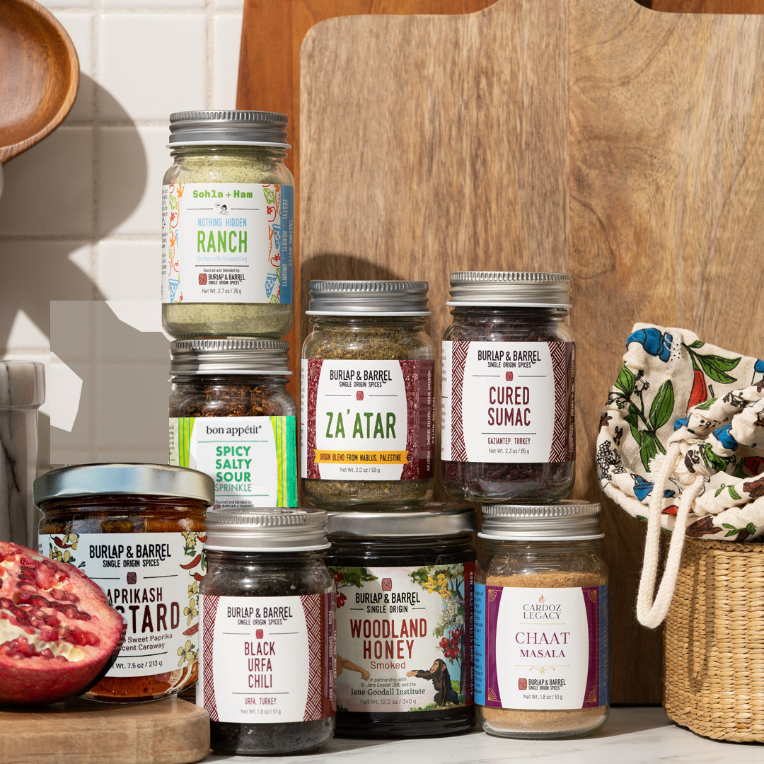 Collection of Burlap & Barrel spice jars on a wooden surface with a pomegranate and floral basket.