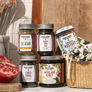 Collection of Burlap & Barrel spice jars on a kitchen counter with a pomegranate and basket in the background.