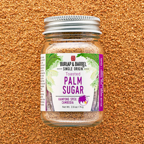 A glass jar of Burlap & Barrel Toasted Palm Sugar, featuring a white and purple label with green palm leaf graphics, sealed with a silver metal lid and set against a textured brown background.