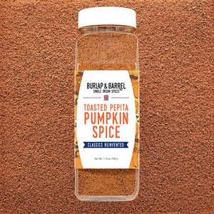 A clear plastic spice jar of Burlap & Barrel Toasted Pepita Pumpkin Spice with a white lid and white label, set against a textured brown background.